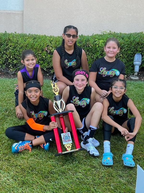 Team Legacy girls' basketball team posing outdoors with a red championship trophy after a successful tournament. The young athletes, dressed in black uniforms with colorful 'Legacy' logos, display a variety of joyful expressions. They are seated on t