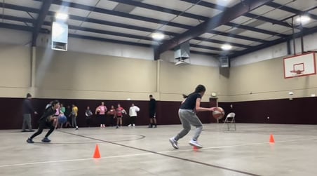 Team Legacy Basketball practice session in progress, with a young athlete dribbling towards an orange cone on an indoor court. The gym is spacious with a high ceiling, brown walls, and a visible basketball hoop in the background. Other team members a