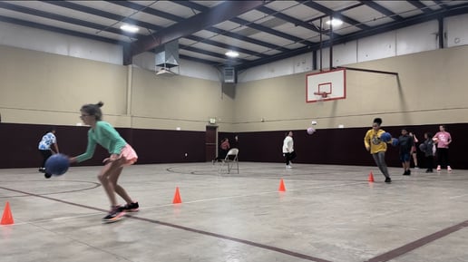 Girls from Team Legacy Basketball practice dribbling and ball-handling skills on an indoor court with a basketball hoop in the background. They are actively engaged in a drill, maneuvering around orange cones. The gym features brown walls and is lit 