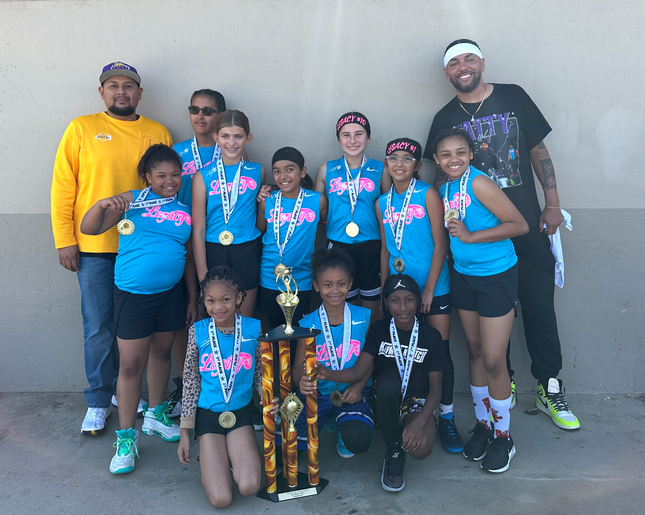 Team Legacy Basketball girls' team posing with medals and a trophy after a successful tournament. The team, in matching blue jerseys with 'Legacy' emblazoned across the front, are smiling proudly alongside their coaches. The backdrop is a plain wall,